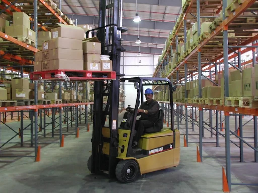 A forklift working between tall pallet racks stacked with cartons inside a warehouse