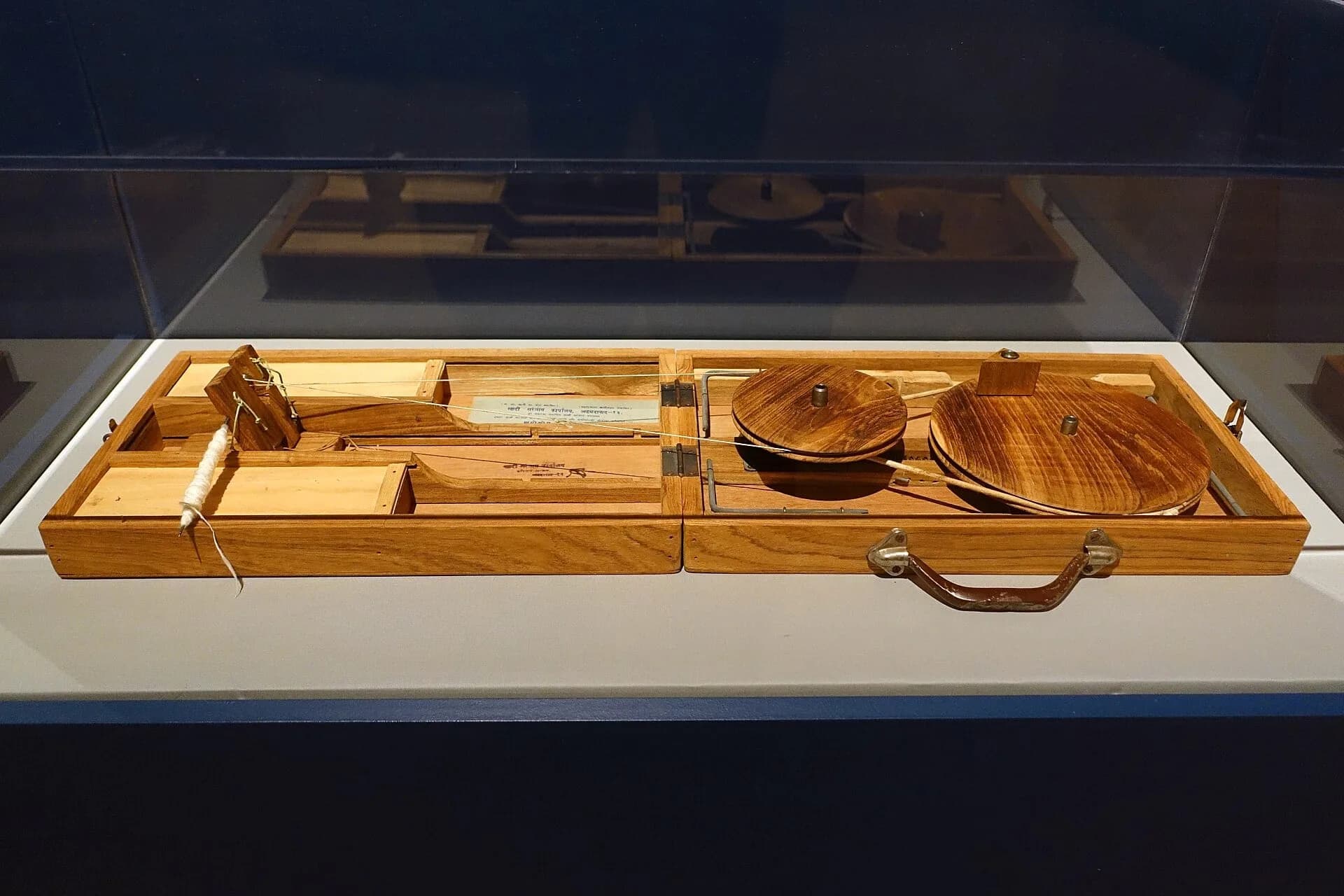 A wooden box charkha, a portable spinning wheel used to spin khadi yarn, displayed in a museum case