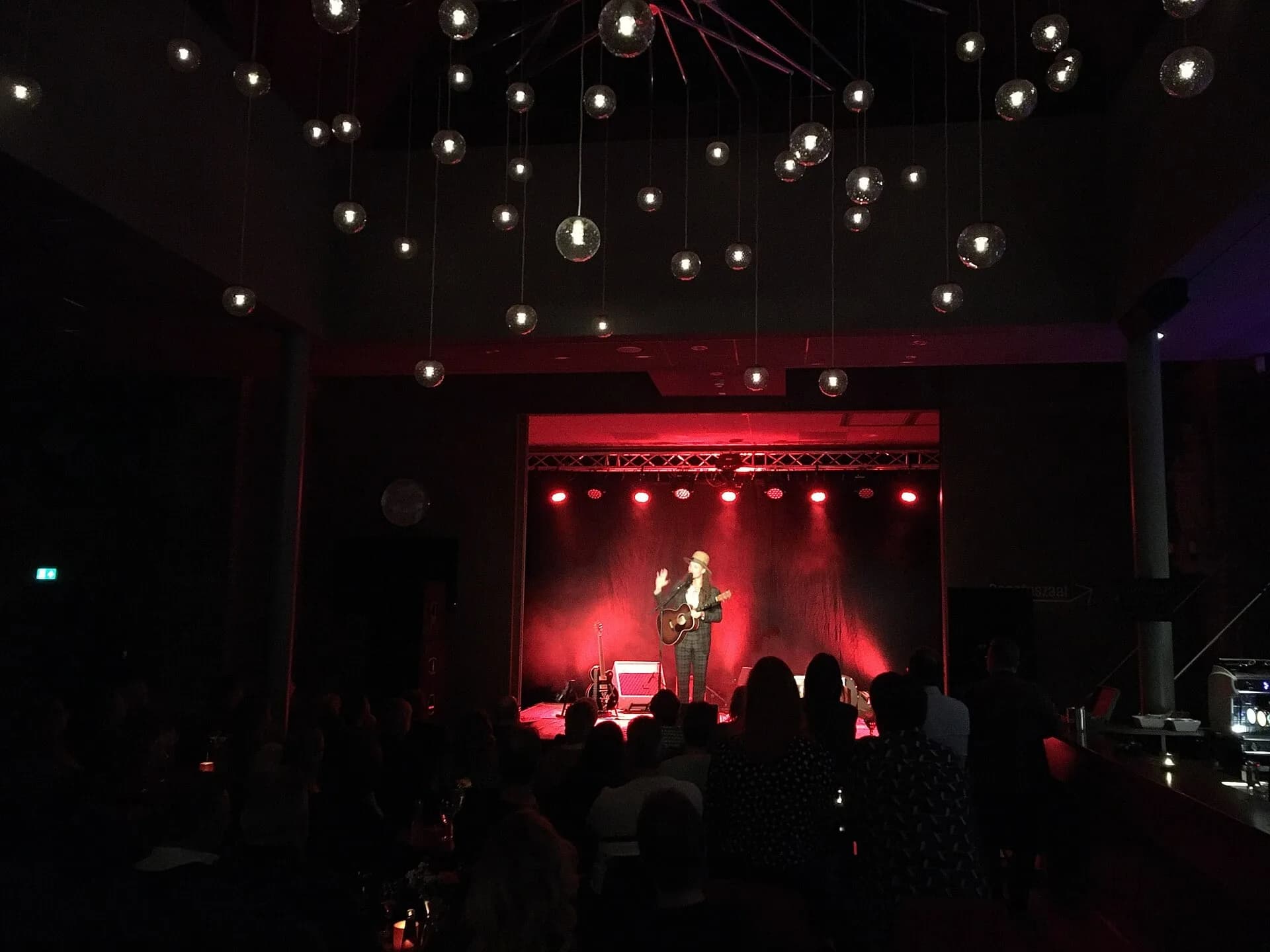 A performer on a small stage lit in red, with strings of lights above a darkened audience