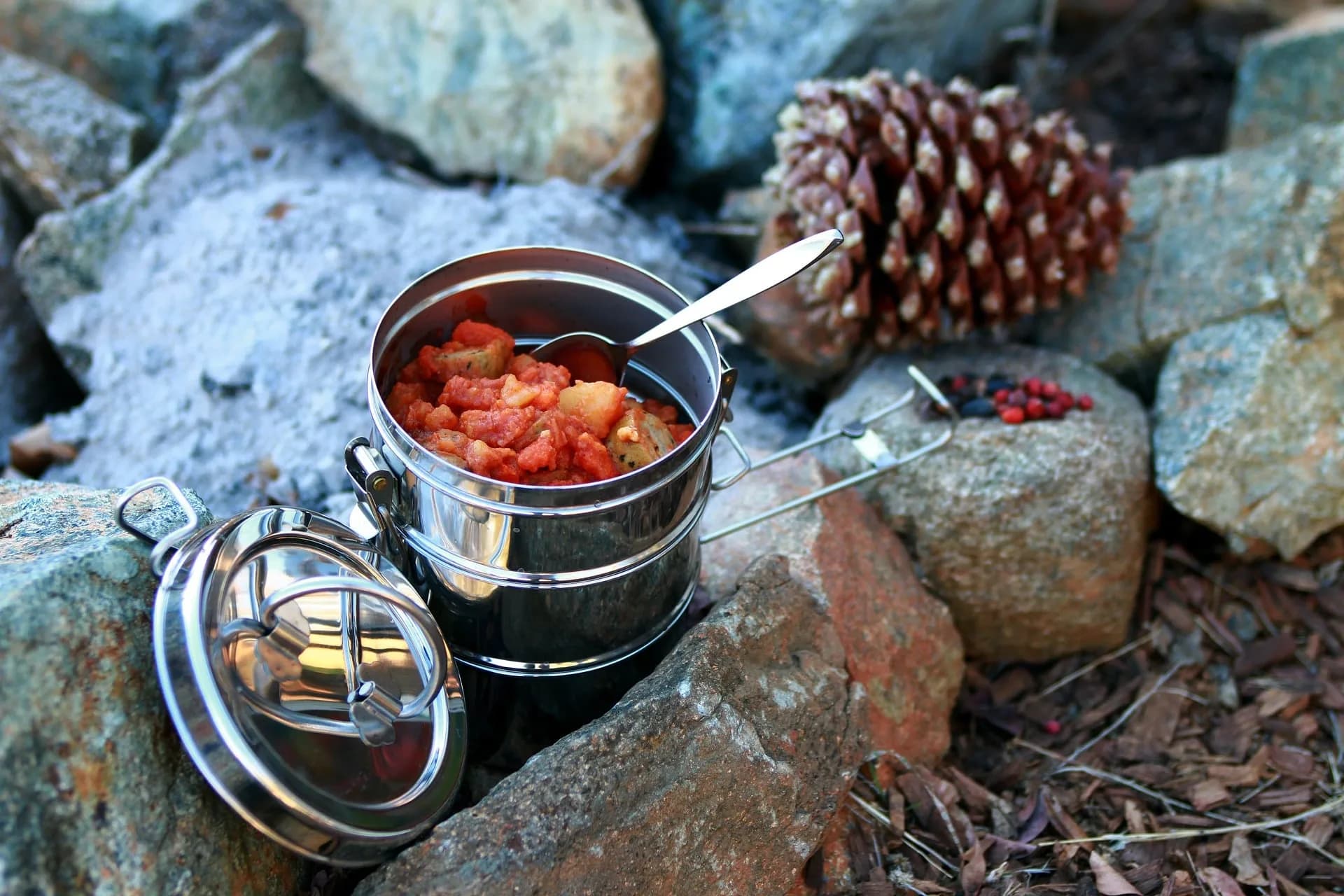 A stacked metal food container with a meal inside, resting on rocks outdoors